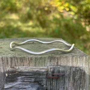 May include: A silver-colored, wavy bangle bracelet. The bracelet has a fluid, organic design, with a smooth, polished finish. The bracelet is resting on a weathered wooden surface, with a blurred green background.