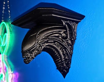 May include: A black shelf in the shape of an Alien Xenomorph head, mounted on a blue wall. A neon sign of an alien head hangs to the left. The shelf has intricate details and a flat top.
