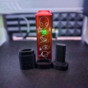 Peut inclure: Un appareil &eacute;lectronique rouge et argent&eacute; avec un design sur le th&egrave;me des extraterrestres et de l'espace, accompagn&eacute; de trois composants noirs. L'appareil a une lumi&egrave;re verte, probablement un produit de vapotage.