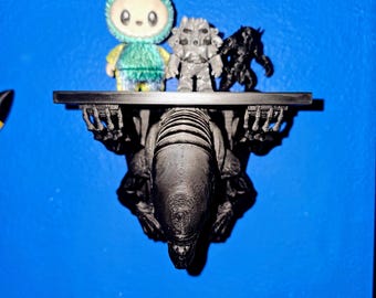 May include: A black shelf with three figurines on it, mounted on a blue wall. The figurines include a green and yellow bunny-like character, a gray robot, and a black monster. The shelf is supported by a large, black alien-like figure.
