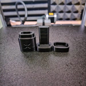 Peut inclure: Trois objets noirs imprim&eacute;s en 3D sont dispos&eacute;s sur une surface textur&eacute;e. Les objets comprennent une base rectangulaire avec un motif en X, une pi&egrave;ce rectangulaire haute et une petite pi&egrave;ce de forme ovale. Il s'agit de composants d'un produit plus grand.