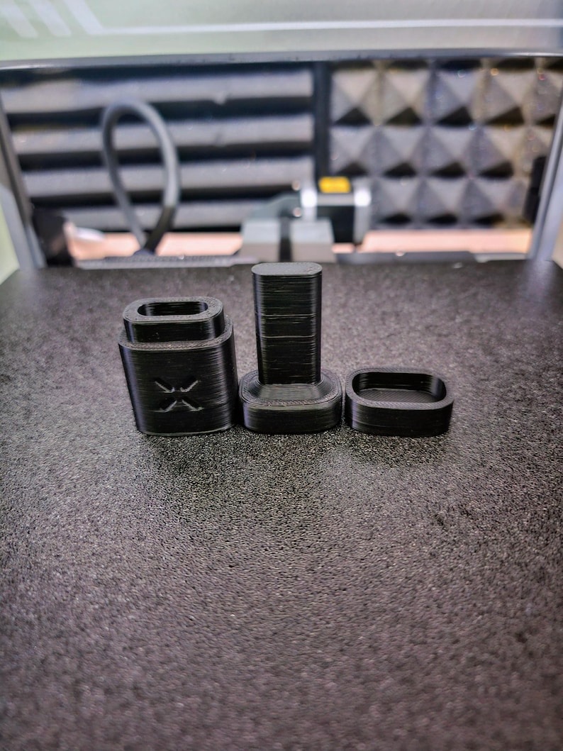 Peut inclure: Trois objets noirs imprim&eacute;s en 3D sont dispos&eacute;s sur une surface textur&eacute;e. Les objets comprennent une base rectangulaire avec un motif en X, une pi&egrave;ce rectangulaire haute et une petite pi&egrave;ce de forme ovale. Il s'agit de composants d'un produit plus grand.