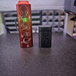 Peut inclure: Un appareil cylindrique rouge avec un design d'extraterrestre et de corps c&eacute;lestes, et un petit &eacute;tui rectangulaire noir. L'appareil rouge a une lumi&egrave;re verte et des symboles grav&eacute;s. L'&eacute;tui noir a un motif sculpt&eacute; sur le devant.