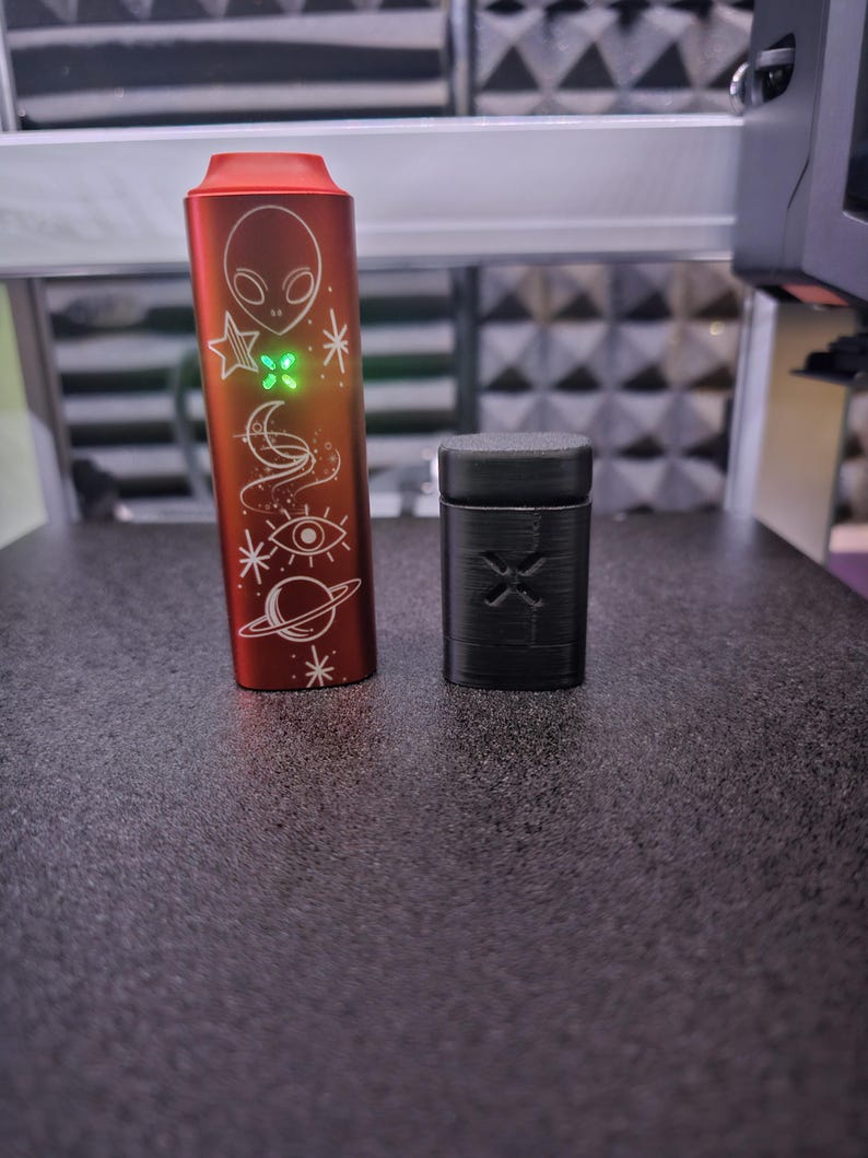 Peut inclure: Un appareil cylindrique rouge avec un design d'extraterrestre et de corps c&eacute;lestes, et un petit &eacute;tui rectangulaire noir. L'appareil rouge a une lumi&egrave;re verte et des symboles grav&eacute;s. L'&eacute;tui noir a un motif sculpt&eacute; sur le devant.