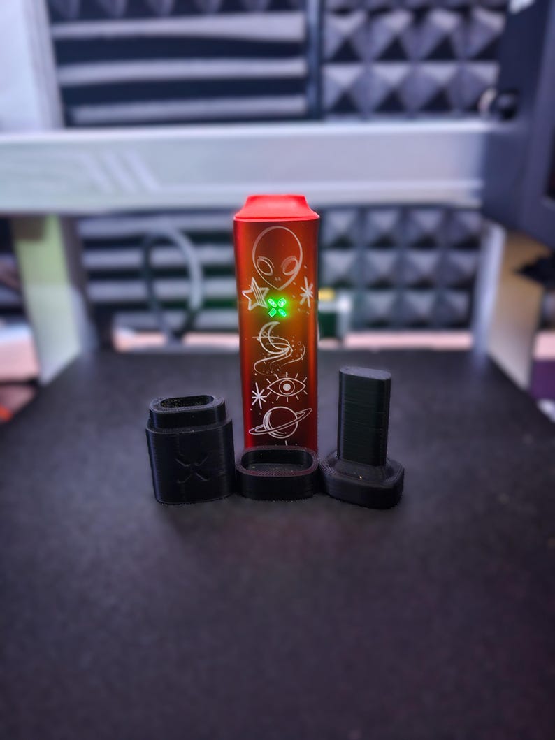 Peut inclure: Un appareil &eacute;lectronique rouge et argent&eacute; avec un design sur le th&egrave;me des extraterrestres et de l'espace, accompagn&eacute; de trois composants noirs. L'appareil a une lumi&egrave;re verte, probablement un produit de vapotage.