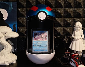 May include: A blue and white spherical display case featuring a central black section holding a Palkia trading card. The top half is blue with red accents, while the bottom half is white. The case is positioned between a white tree sculpture and a white figurine.