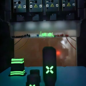 May include: Several black objects with glowing green details, including a rectangular shape, two small cubes, and a tall, cylindrical item. They are on a blue surface, with a complex machine above.
