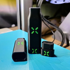 May include: Three black vaping device components with green cross-shaped accents. The components include a rectangular mouthpiece, a tall cylindrical body, and a small rectangular piece. The items are on a blue surface.