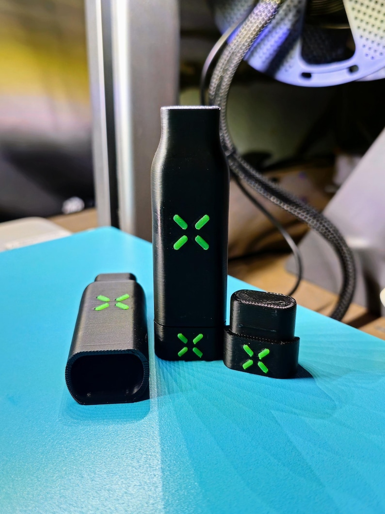 May include: Three black vaping device components with green cross-shaped accents. The components include a rectangular mouthpiece, a tall cylindrical body, and a small rectangular piece. The items are on a blue surface.