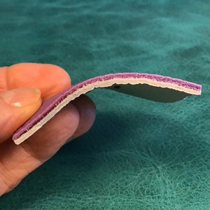 Puede incluir: Primer plano del lado de un material de cuero morado con una capa blanca debajo. El material es flexible y tiene una superficie ligeramente texturizada.