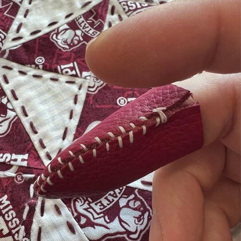 May include: A close-up of a burgundy leather thimble with white stitching. The thimble is worn on a finger and is being used for sewing.