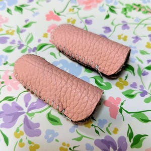 Könnte beinhalten: Zwei rosafarbene Fingerprotektoren aus Leder mit sichtbaren Nähten. Die Fingerschützer bestehen aus strukturiertem Leder und sollen die Finger schützen. Der Hintergrund ist ein Blumenmuster mit rosa, lila und grünen Blumen.