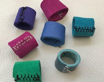 May include: A collection of colorful leather finger protectors in shades of purple, pink, blue, green, and teal. Each finger protector is cylindrical and features contrasting stitching in black or white thread. The items are arranged on a white surface.