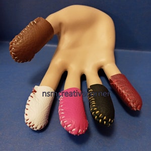 May include: Five leather thimbles in different colors, including brown, white, pink, black, and red. Each thimble has white stitching around the edge.