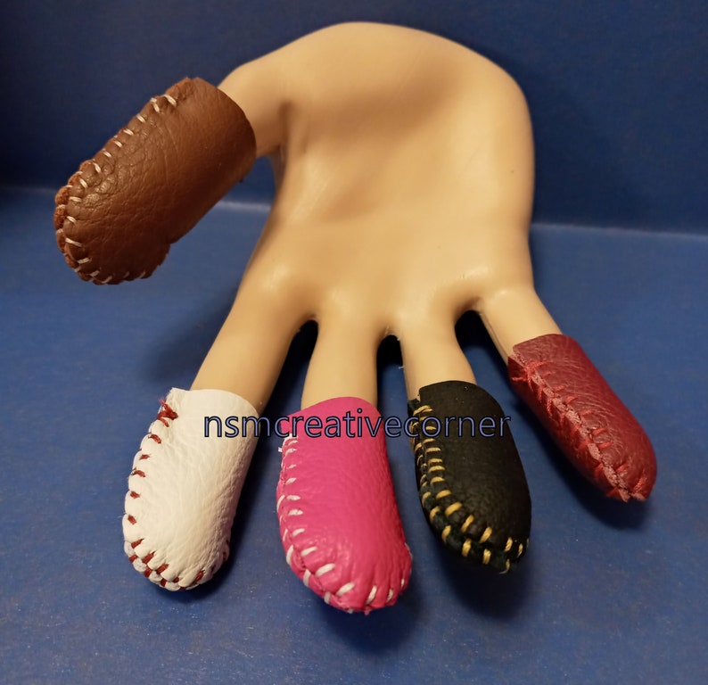 May include: Five leather thimbles in different colors, including brown, white, pink, black, and red. Each thimble has white stitching around the edge.