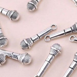 Könnte beinhalten: Silberfarbene Mikrofon-Anhänger auf einer hellrosa Oberfläche. Die Charms haben einen detaillierten Mikrofonkopf und eine Schlaufe zum Befestigen an Schmuck oder anderen Bastelarbeiten. Ideal für Musik-Themenprojekte.