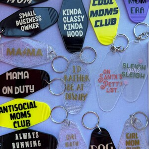 以下が含まれることがあります： さまざまな形と色のノベルティキーホルダーのコレクション。「Cool Mom」、「Dog Mom」、「I'd Rather Be at Disney」などのフレーズが特徴です。キーホルダーはアクリルとプラスチック製で、金属製のキーリングが付いています。