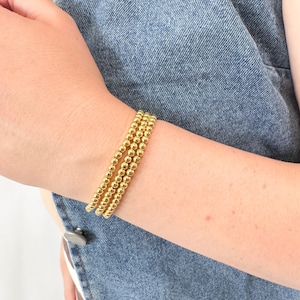 May include: Three gold beaded bracelets are stacked on a wrist. The bracelets are made of small, round, gold beads. The person is wearing a denim jacket. The bracelets are a simple and elegant accessory.