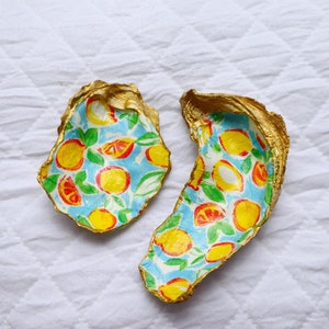 Op de afbeelding: Twee oesterschelpen met een gouden rand en een beschilderd interieur. Het interieur heeft een blauwe achtergrond met gele citroenen en sinaasappelschijfjes. De schelpen zijn gerangschikt op een wit, gewatteerd oppervlak.