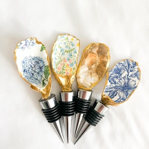 May include: Four decorative bottle stoppers with unique designs. Each stopper features a different design on an oyster shell, including blue floral patterns and a natural shell appearance. The stoppers have silver metal bases and black rubber seals.