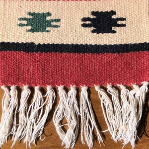 May include: A red, white, and black woven rug with a fringe border. The rug has a geometric pattern with green and black shapes.