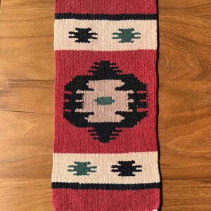 May include: A red, white, and black woven rug with a geometric pattern. The rug has a central design with a green circle and a smaller green square in the center. The rug is approximately 18 inches long and 6 inches wide.