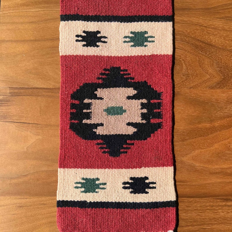 May include: A red, white, and black woven rug with a geometric pattern. The rug has a central design with a green circle and a smaller green square in the center. The rug is approximately 18 inches long and 6 inches wide.
