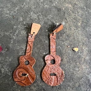 May include: A pair of brown leather guitar-shaped earrings with a wood top. The earrings have a textured surface and a circular sound hole. The earrings are on a dark gray surface.