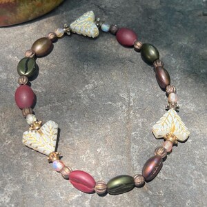 Puede incluir: Una pulsera de cuentas con cuentas burdeos, verde oliva y bronce. La pulsera incluye cuentas en forma de corazón con un diseño de hoja dorada. Las cuentas están ensartadas con una delicada cadena. Longitud: aprox. 18 cm.