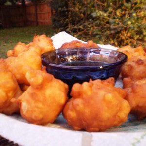 Könnte beinhalten: Ein Teller mit goldbraunen, frittierten Krapfen mit einer dunkelblauen Glasschale mit Sirup in der Mitte. Die Krapfen sind auf einem weißen Tuch gestapelt, mit einem verschwommenen grünen Hintergrund aus Gras und Laub.