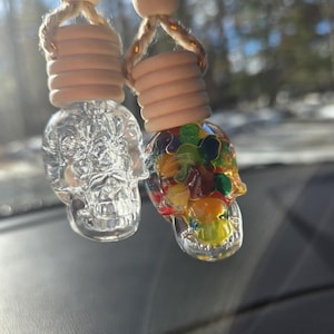 Può includere: Due profumatori per auto a forma di teschio appesi a un filo. Uno è in vetro trasparente, l'altro è pieno di perline colorate. Ognuno ha una perlina di legno e un anello di legno attorno alla parte superiore. Lo sfondo è sfocato.