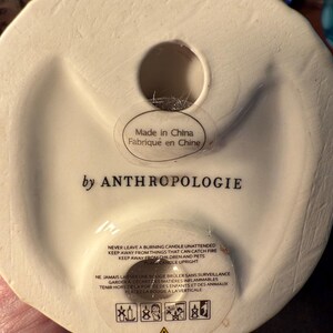 May include: Off-white ceramic candle holder base with a circular opening. The text "Made in China" and "by ANTHROPOLOGIE" are visible. Safety instructions in English and French are printed on the base, along with warning symbols.