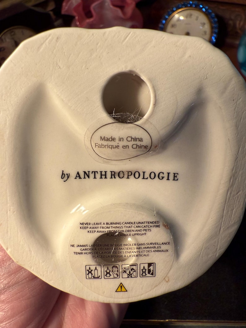 May include: Off-white ceramic candle holder base with a circular opening. The text "Made in China" and "by ANTHROPOLOGIE" are visible. Safety instructions in English and French are printed on the base, along with warning symbols.