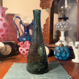 May include: A tall, dark green glass bottle with a crackle finish. The bottle is displayed on a vintage book with a green cover. Other antique glass objects are in the background.