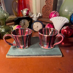 May include: Two ceramic mugs with a pink, brown, and green striped design. The mugs are sitting on a green book with a brown cover.