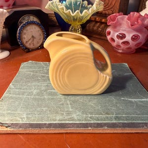 May include: A vintage, light yellow ceramic pitcher with a curved handle and a circular design. The pitcher has a small spout and a blue accent on the handle. It is sitting on a green surface.