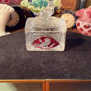 May include: A clear glass trinket box with a bird-shaped lid. The box has a red and clear bird design on the front. The box is sitting on a dark surface, with other decorative items in the background.