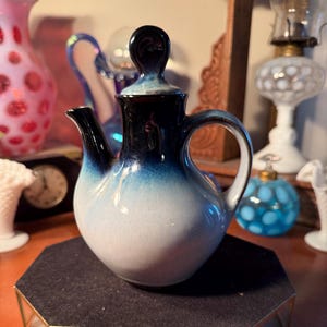 May include: A small, ceramic teapot with a handle and spout. The teapot is a gradient of colors, transitioning from dark blue at the top to white at the bottom. The lid has a rounded handle. The teapot is displayed on a black surface.