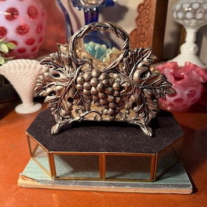 May include: A silver-toned metal basket with a handle, adorned with grape and leaf motifs. The basket sits atop a black velvet-covered base, which is set on a multi-faceted, glass-paneled display stand. The stand has a gold trim and a light green base.