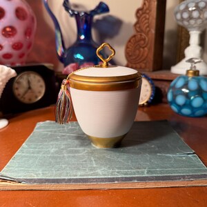 May include: A white and gold lidded container with a tassel, resting on a green book. The container has a gold handle and trim. Other decorative items, including a clock and vases, are in the background.
