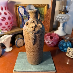 May include: A tall, cylindrical stoneware jug with a speckled tan and brown finish. The vessel features a sculpted, relief-style face on the upper body, depicting a figure with a prominent mustache, a pointed beard, and two small, rounded ears.