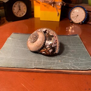 May include: A decorative seashell with a large, white, iridescent center surrounded by a ring of rhinestones. The shell is brown and black with a pearlescent sheen.