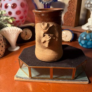 May include: A handmade, rustic ceramic mug with a cowboy face design. The mug features a brown rim and a tan body with a sculpted face wearing a cowboy hat. The mug is displayed on a black and glass display stand.