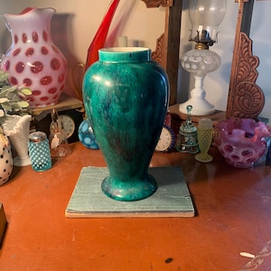 May include: A tall, green vase with a speckled pattern. The vase is placed on a green book and sits on a brown table. Other decorative items, including a pink vase with red spots, are visible in the background.