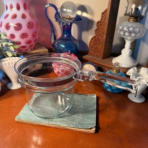 May include: A clear glass saucepan with a handle sits on a small, aged, green book. The saucepan is round with a clear handle. The background features various antique glass vases and lamps in shades of pink, blue, and white.