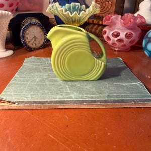 May include: A lime green ceramic pitcher with a circular design. The pitcher has a handle and a spout. The pitcher is sitting on a green surface. Other vintage glassware is in the background.