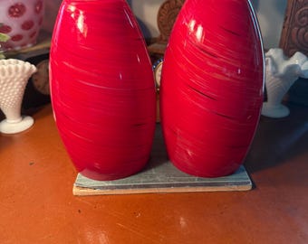 May include: Two red glass vases with a swirl design. The vases are oval-shaped and sit on a small, rectangular base. The vases have a glossy finish and are displayed on a brown surface.