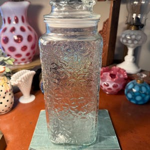 May include: A clear glass jar with a textured floral pattern, topped with a matching lid. The jar is rectangular and sits on a light blue book. Other decorative glass items are in the background.