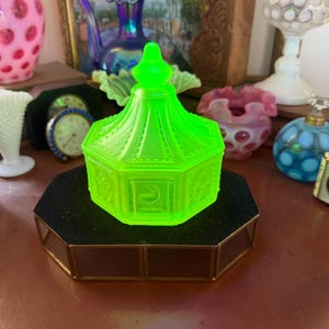 May include: A bright neon green glass candy dish with a lid, featuring an octagonal shape and a pointed finial. Each side of the dish displays a distinct embossed panel, including images of a bird, floral motifs, and other decorative patterns.