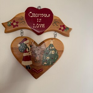 May include: Wooden Christmas decoration with a heart-shaped design. The top heart reads "Christmas is Love" in white lettering on a red background. The bottom heart features Santa Claus, a Christmas tree, and other festive elements.